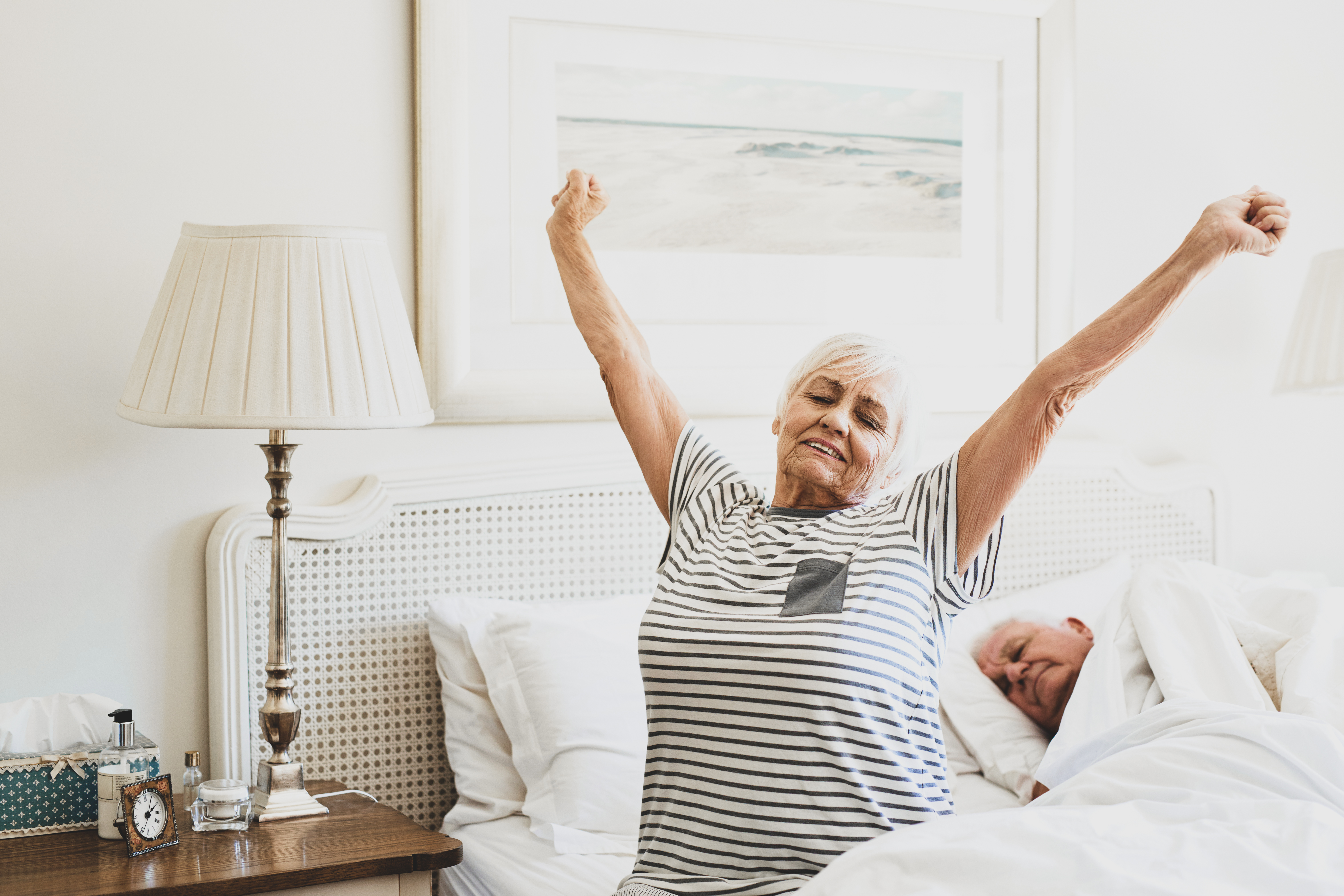 Senior woman sitting on her bed in the morning yawning with arms raised in a stretch with her husband sleeping next to her