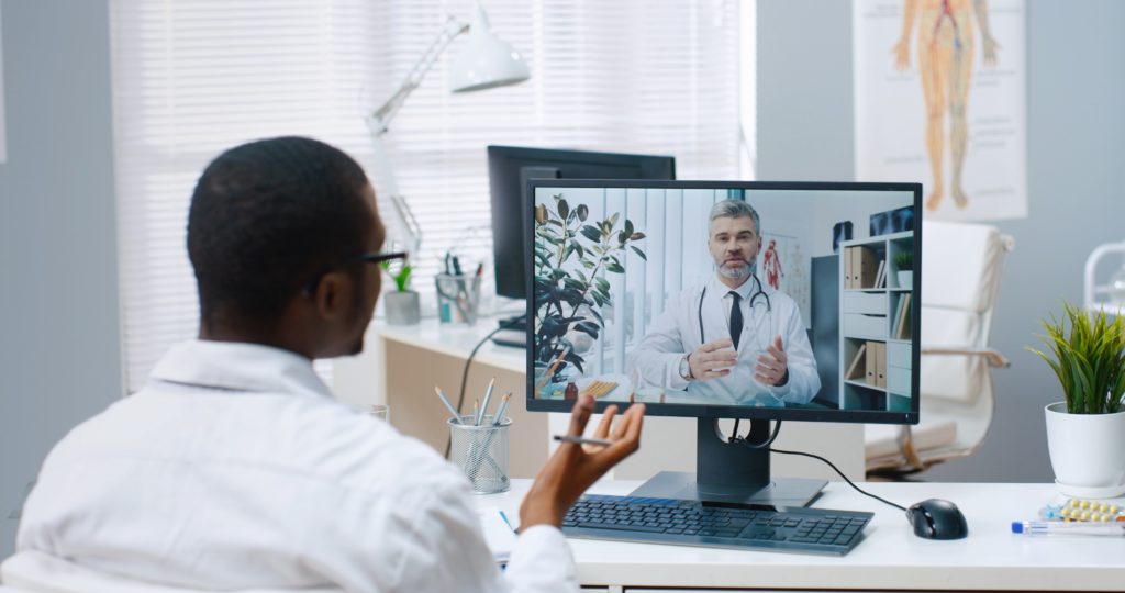 Over shoulder view of African American male specialist talks on online video call with Caucasian middle-aged doctor on computer sitting in office experienced physician consults nurse on web conference