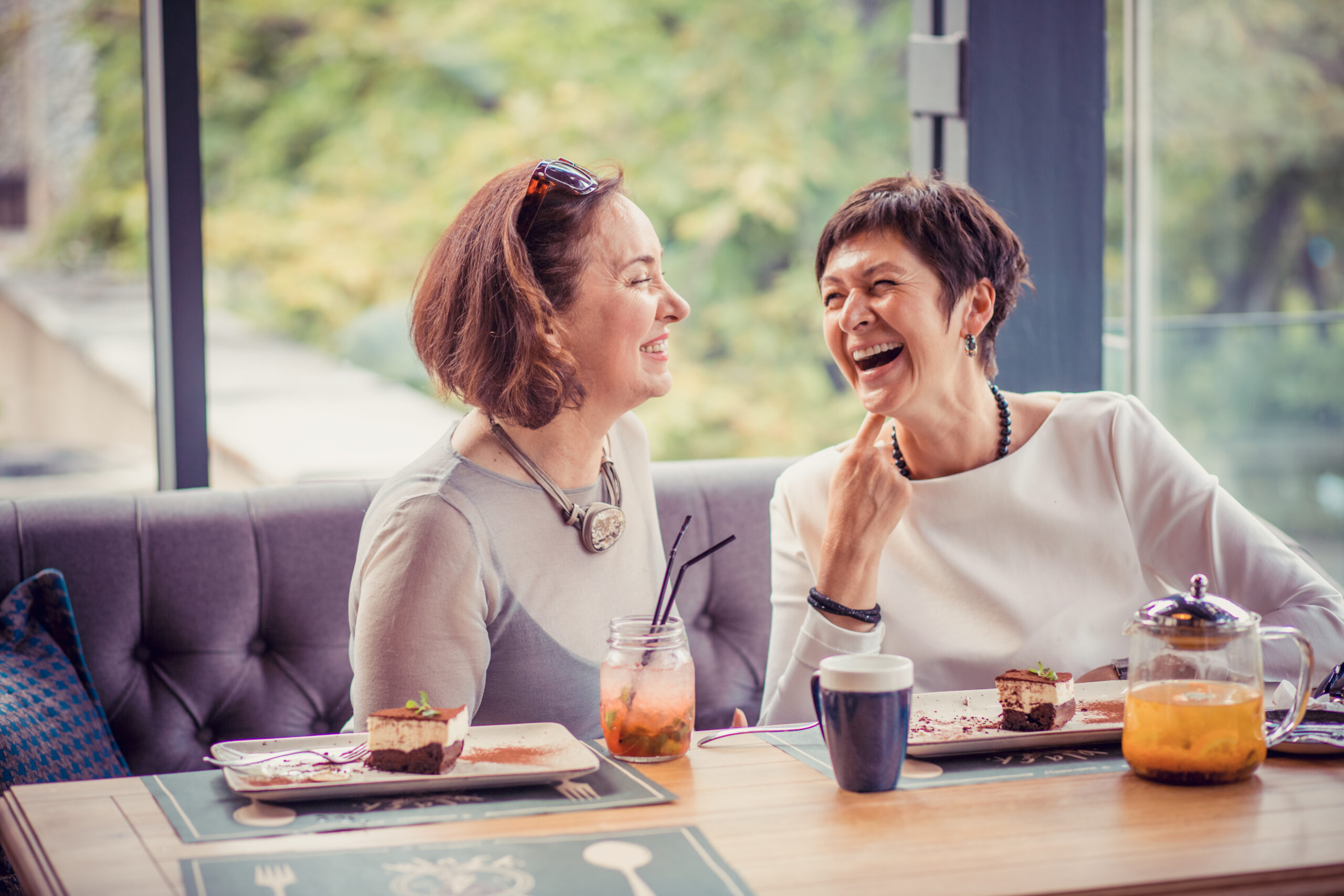 Beautiful trendy middle-aged women having coffee with desserts at table in cafe and laughing in leisure