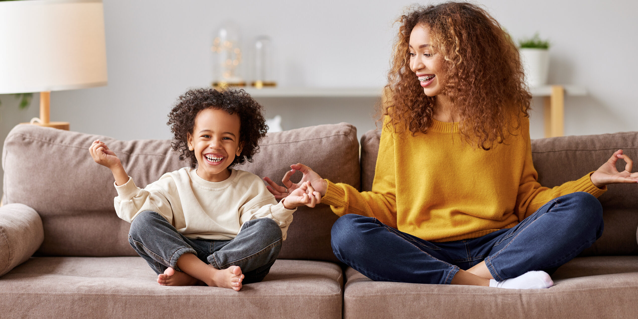 Yoga with kids. Young afro american family mother and little son meditating while sitting in lotus pose on sofa in living room, smiling and enjoying leisure time together at home