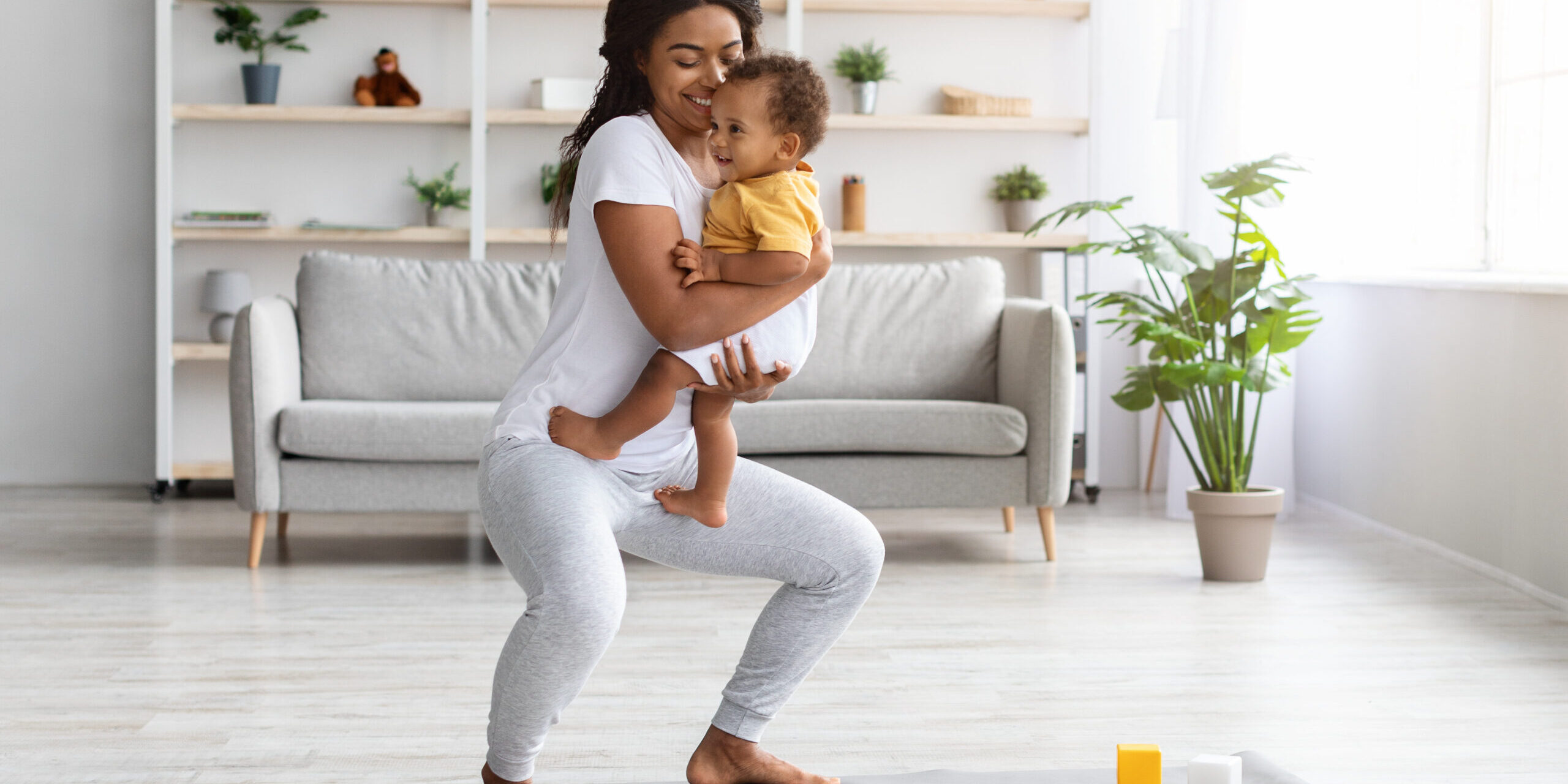 Postpartum Weight Loss. Happy Black Lady Making Squats With Baby On Hands While Training At Home, Smiling African American Woman Exercising In Living Room And Bonding With Infant Child, Copy Space