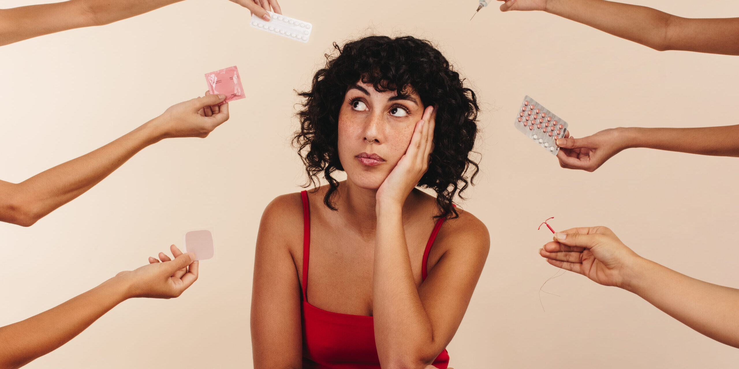 Young woman looking away thoughtfully while surrounded by hands holding different forms of hormonal and non-hormonal contraception. Pensive young woman making a decision about her reproductive health.